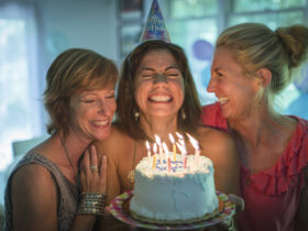Zadowolona kobieta trzyma tort urodzinowy. Obok niej śmieją się dwie koleżanki.