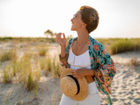 Kobieta na plaży z narzutką w kwiatki. Trzyma w ręku słomiany kapelusz.